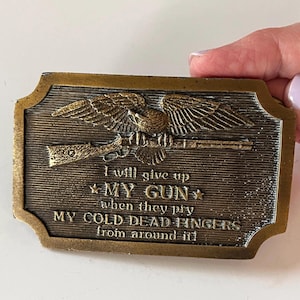 May include: A rectangular brass-toned belt buckle featuring a relief of an eagle perched on a rifle. The text reads: I will give up MY GUN when they pry MY COLD DEAD FINGERS from around it! The background has a horizontal lined texture.