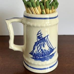 May include: A cream-colored ceramic mug with a blue ship illustration and blue horizontal bands, featuring a textured crackle glaze finish. The mug is filled with a collection of wooden matches that have green tips, creating a functional display.