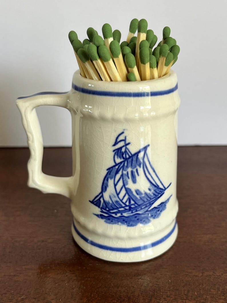 May include: A cream-colored ceramic mug with a blue ship illustration and blue horizontal bands, featuring a textured crackle glaze finish. The mug is filled with a collection of wooden matches that have green tips, creating a functional display.