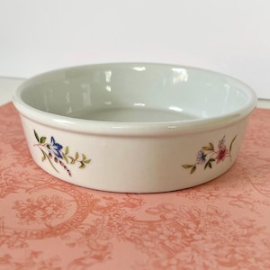 May include: A round, white ceramic bowl with a smooth, glossy finish. The exterior features two delicate, hand-painted floral motifs on opposite sides, showcasing small blue, pink, and green leaves and petals against the plain white surface.