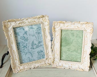 May include: Two rectangular picture frames with ornate, cream-colored borders featuring gold-toned highlights. The left frame contains a light blue insert with a floral and bird pattern, while the right frame holds a light green insert with a similar design.