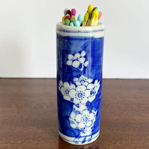 May include: A cylindrical ceramic match holder featuring a blue and white floral pattern. The holder is filled with a collection of wooden matches that have colorful tips in shades of yellow, pink, light blue, and green, protruding from the top.
