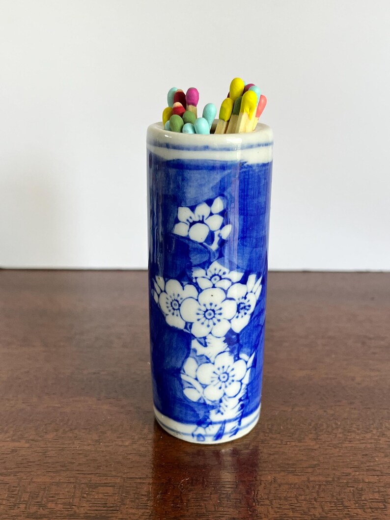 May include: A cylindrical ceramic match holder featuring a blue and white floral pattern. The holder is filled with a collection of wooden matches that have colorful tips in shades of yellow, pink, light blue, and green, protruding from the top.