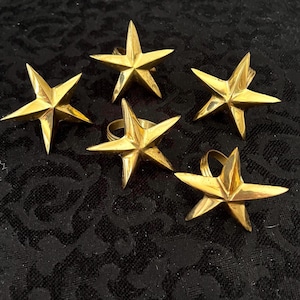 May include: Five gold-colored star-shaped napkin rings. Each ring features a five-pointed star design with a circular ring attached to the back. The napkin rings are made of a shiny, metallic material and are arranged on a dark, textured surface.