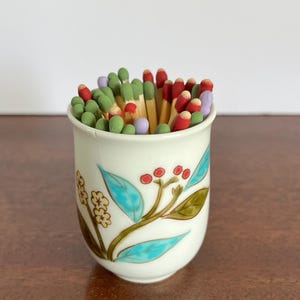 May include: A white ceramic cup filled with wooden matches that have colorful heads in shades of red, green, and light purple. The exterior of the cup features a painted botanical design with turquoise leaves, small red berries, and yellow flowers.