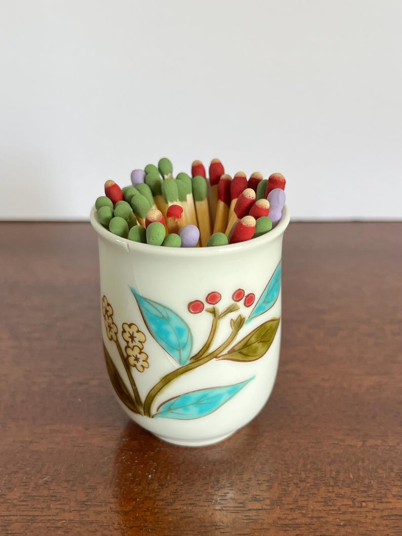 May include: A white ceramic cup filled with wooden matches that have colorful heads in shades of red, green, and light purple. The exterior of the cup features a painted botanical design with turquoise leaves, small red berries, and yellow flowers.