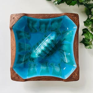 May include: A square ceramic bowl featuring a bright turquoise interior with a dark teal drip glaze pattern. The rim is textured to resemble brown wood grain, creating a contrast between the smooth, glossy center and the rustic border.