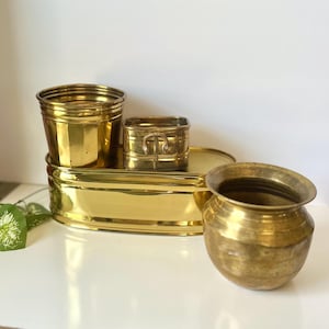 May include: A collection of four polished brass vessels including a large oval basin, a tall cylindrical pot, a small square container with side handles, and a rounded bowl with a flared rim, all featuring a reflective metallic gold finish.