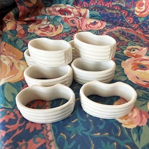May include: A collection of white ceramic napkin rings. Each ring has a rounded shape with a ribbed texture. The napkin rings are arranged on a floral patterned fabric with pink, blue, and green hues.