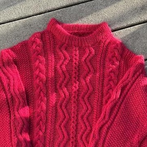 May include: A close-up of a vibrant red knitted sweater featuring a mock neck and cable knit design. The sweater has a textured appearance and is made of a soft, warm material. The cable knit pattern runs down the front and sleeves.