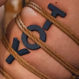 Puede incluir: Primer plano de piel con las letras "XO" en azul oscuro, rodeadas de cuerda marrón. Las letras son tridimensionales y texturizadas. La cuerda está enrollada alrededor de la piel, creando un patrón. Tema sensual.