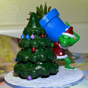 May include: A whimsical Christmas-themed figurine featuring a green Christmas tree decorated with colorful ornaments and a frog wearing a Santa hat and coat, carrying a blue flower pot with a succulent plant. The figurine is on a round, snow-covered base.