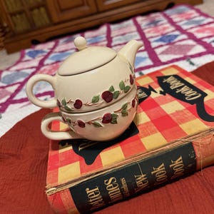 May include: A cream-colored ceramic teapot and teacup set with a floral design of red berries and green leaves. The set is stacked on top of a vintage cookbook with a red and yellow checkered pattern.