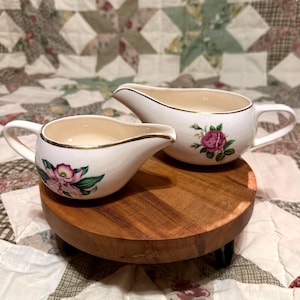 May include: Two white ceramic cream pitchers with gold trim and floral designs. One pitcher features a pink and green orchid, while the other has a pink rose. The pitchers are on a round wooden stand.