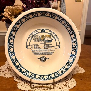 May include: A round, cream-colored ceramic bowl featuring a dark blue decorative border with a repeating arch pattern. The center displays a black graphic illustration of Caesar salad ingredients and a printed recipe for Caesar salad.