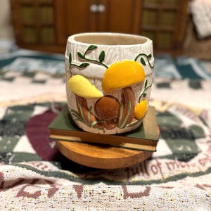 May include: A ceramic mug with a mushroom design. The mug is cylindrical with a cream-colored background and a textured, wood-like appearance. Yellow, brown, and orange mushrooms are painted on the mug, with green vines. The mug is sitting on a stack of books.