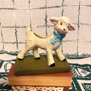 May include: A ceramic lamb figurine planter with a textured cream-colored coat, pink-tinted cheeks, and a bright blue bow tied around its neck. The lamb has black-painted hooves and is standing on top of two stacked hardcover books.