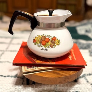 May include: White enamel tea kettle with a black handle and lid. The kettle features a floral design with peaches and the text "Le Thé". The kettle sits on top of a stack of books.