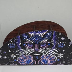 Può includere: Un oggetto decorativo nero a forma di mezza luna con un design a forma di gatto. Il volto del gatto è adornato con strass blu, viola e bianchi, creando un effetto scintillante. Lo sfondo è nero con stelle bianche e accenti floreali.
