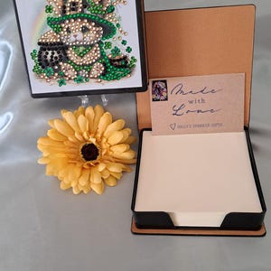 May include: A black and brown notepad holder with a St. Patrick's Day theme. The image shows a bunny wearing a green hat and holding a pot of gold. A yellow flower is next to the notepad holder. The notepad holder has a card that says "Made with Love".