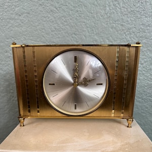 May include: A gold-toned rectangular clock with a round face. The clock face is silver with black lines marking the hours. The clock has decorative gold accents and small feet. The hands are ornate and gold.