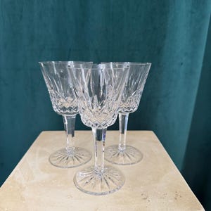 May include: Three clear crystal wine glasses with intricate cut patterns. The glasses have a stemmed base and a flared bowl. They are arranged on a light-colored surface against a dark green backdrop. The glasses are likely for formal dining or special occasions.