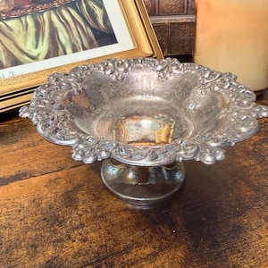 May include: A silver-colored, ornate pedestal bowl with a scalloped edge. The bowl sits on a short, round base and features detailed floral and scrollwork designs. The bowl is displayed on a dark wooden surface.