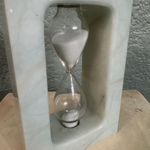 May include: A light green marble hourglass with a clear glass sand timer. The hourglass is set within a rectangular marble frame. The sand is white, and the marble has a natural veining pattern.