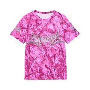 May include: A pink camouflage pattern t-shirt with short sleeves. The shirt has silver text across the chest and on the sleeves. The text is a stylized band logo. The t-shirt has a crew neck.