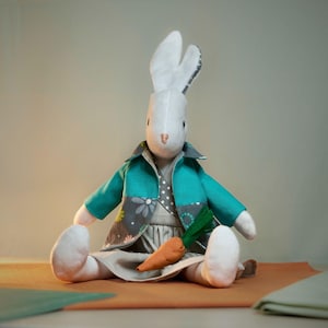 May include: A white stuffed bunny wearing a teal jacket with gray floral accents and a gray dress. The bunny is holding an orange carrot. The bunny has long white ears with gray patterned lining.