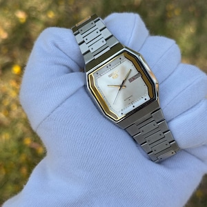 Könnte beinhalten: Silberfarbene Seiko 5 Automatik-Uhr mit rechteckigem Zifferblatt und goldfarbenen Akzenten. Das Zifferblatt ist weiß mit schwarzen Zeigern und Markierungen, Tag und Datum werden angezeigt. Das Armband besteht aus silberfarbenen Metallgliedern.