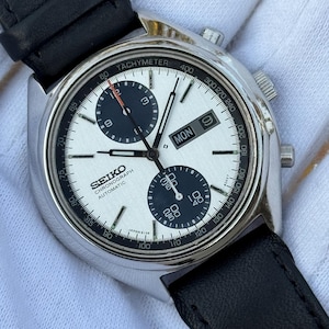 May include: A silver Seiko Chronograph automatic watch with a white face, black sub-dials, and a black leather band. The watch face displays the day and date. The watch has a tachymeter scale and is a classic timepiece.