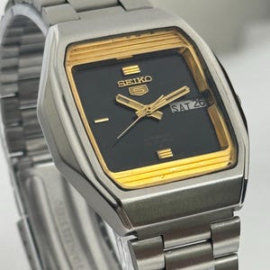 Puede incluir: Un reloj de pulsera automático Seiko 5 plateado con esfera negra y detalles dorados. El reloj muestra el día y la fecha, con "SAT 26" visible. La correa es de acero inoxidable.