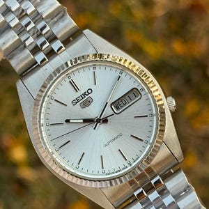 Op de afbeelding: Zilveren Seiko 5 automatische horloge met een gegroefde lunette en een zilveren wijzerplaat. Het horloge heeft een dag- en datumweergave en een roestvrijstalen armband. Het horloge wordt getoond tegen een vage achtergrond.