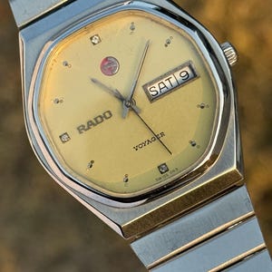 May include: A vintage Rado Voyager watch with a gold-toned face and silver and gold-toned metal strap. The watch face displays the day and date, with the word "Rado" and "Voyager" visible. The watch has a Swiss 063 marking.