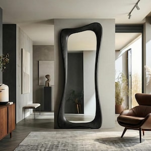 Puede incluir: Un espejo grande de madera teñida oscura con una forma orgánica y fluida. El espejo es alto y estrecho, con un marco curvo. El espejo está colocado en un espacio de vida moderno con un sillón de cuero marrón y una alfombra gris.