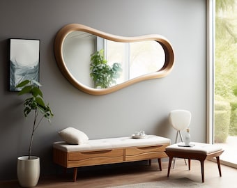 May include: A large, organically shaped wooden mirror with a light-colored frame hangs on a gray wall. Below, a wooden bench with a cream-colored cushion and two drawers sits. A small stool and a potted plant are also in the room.