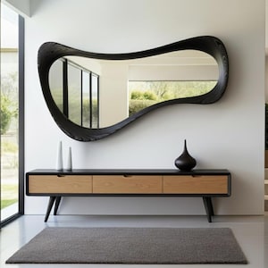 May include: A contemporary mirror with a black, undulating frame is mounted above a wooden console table. The console features three drawers with light wood fronts and a dark frame. Two white vases and a dark vase are on the console.