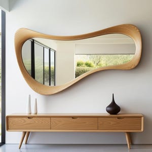 Op de afbeelding: Een grote, organisch gevormde houten spiegel hangt boven een houten console. De spiegel heeft een vloeiend, golvend ontwerp. De console heeft drie lades en taps toelopende poten. Twee witte vazen en een donkerbruine vaas staan op de console.