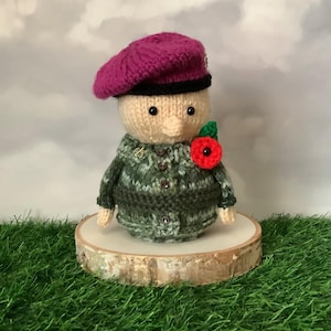 May include: A knitted doll wearing a magenta beret, a green camouflage jacket, and a red poppy. The doll is displayed on a wooden slice. The background features grass and a cloudy sky. The doll is a handmade item.