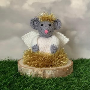 Peut inclure: Figurine d'ange souris tricotée à la main. La souris a des oreilles grises, un nez rose et des yeux noirs. Elle porte un haut blanc tricoté avec des ailes blanches et est entourée de guirlandes dorées. La figurine est posée sur une tranche de bois.