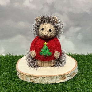 Peut inclure: Figurine de hérisson tricoté à la main, vêtu d'un pull rouge orné d'un sapin de Noël vert. Le hérisson a des piquants bruns et argentés et est posé sur une tranche de bois. Le fond est un ciel nuageux.