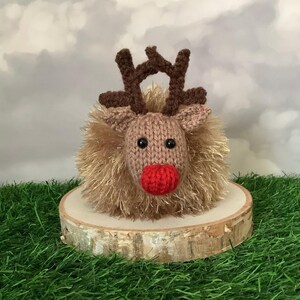 May include: A knitted reindeer ornament with a red nose, brown antlers, and black eyes. The reindeer has a fluffy, light brown body and sits on a wooden slice. The background shows a cloudy sky and green grass.