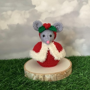 Puede incluir: Figura de ratón tejido, vestida con un atuendo navideño rojo y blanco, que incluye una capa y una diadema con un corazón rojo y hojas verdes. El ratón tiene orejas grises y una nariz rosa, de pie sobre una rodaja de madera.