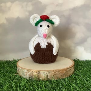 May include: A knitted mouse ornament, featuring a white head and body atop a brown base, adorned with a red and green floral crown. The mouse has a pink tongue and is presented on a wooden slice, against a cloudy background.