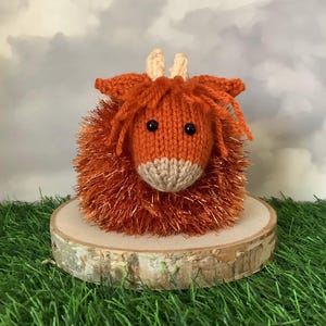 Peut inclure: Figurine de vache des Highlands tricotée à la main, orange et beige, avec des yeux noirs et de petites cornes beiges. Le corps de la vache est duveteux, orange et scintillant. Elle est posée sur une tranche de bois. Fond avec ciel nuageux et herbe verte.