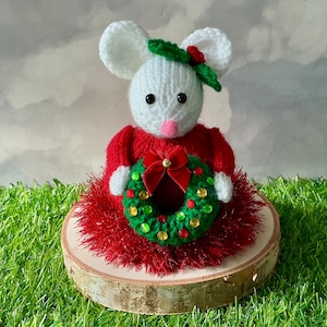 Peut inclure: Une figurine de souris crochetée blanche portant une robe tricotée rouge avec une jupe duveteuse. La souris a un nez rose, des yeux noirs et un accessoire en forme de feuille verte sur la tête. Elle tient une petite couronne verte ornée de perles.