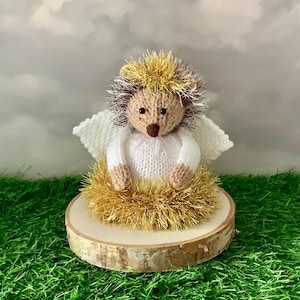 Peut inclure: Figurine d'ange hérisson tricotée à la main. Le hérisson a un visage beige, des yeux bruns et un halo doré. Il porte une robe blanche tricotée et a des ailes blanches. La figurine est posée sur une base en tranche de bois, entourée de tinsel doré.