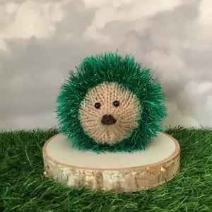 Puede incluir: Un juguete de erizo tejido a mano con una cara beige, nariz marrón y ojos negros. Las púas del erizo están hechas de hilo verde brillante. El juguete se exhibe sobre una pequeña rodaja de madera, sobre un fondo de hierba verde y un cielo nublado.