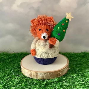 Peut inclure: Figurine de renard tricoté à la main, avec une fourrure orange et un pull blanc et bleu, tenant un sapin de Noël vert orné de décorations colorées et d'une étoile. Le renard est présenté sur une tranche de bois.